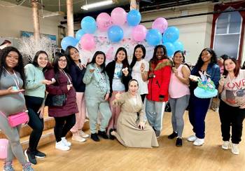 A large group including many pregnant women stands in front of a decorative backdrop with blue and pink balloons.